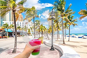 Seafront beach promenade with hand holding cocktail and palm trees on background on sunny day in Fort Lauderdale in