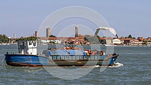 A seagull flies over an old ship with a crane in the lagoon of Venice, Italy, with the island of Murano in the background
