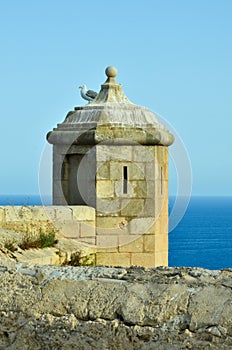 A seagull sits on a castle tower of castillo de Santa BÃ¡rbara in Alicante overlooking the Mediterranean Sea under clear blue sky