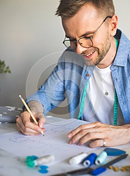 Seamster with glasses is working at the table.DIY designer draws a sketch for a new costume project.Smiling man in a blue shirt