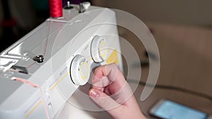 The seamstress chooses the mode on the sewing machine. She rotates the switch with her hand. The camera slides to the