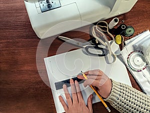 Seamstress draws a pattern with a pencil and a ruler on a white sheet of paper against the sewing supplies. Top view on a brown