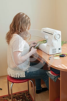 Seamstress sitting on blue chair near wooden table with white sewing machine and holds scissors