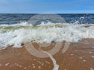 Seascape of the Baltic sea with big foamy waves with yellow sand on the shore and clear blue sky above