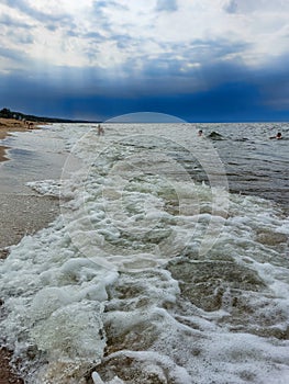 Seascape of the Baltic sea with big foamy waves with yellow sand on the shore and very dark, blue, contrasting clouds above