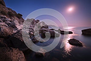 Seascape with moon and lunar path with rocks at night