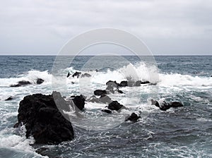 Seascape with stormy dramatic waves breaking over coastal rocks with white surf and grey sky