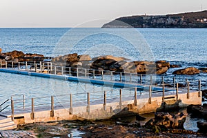 Seaside ocean swimming pool with metal hand rail fencing set against calm blue water and large exposed rocks