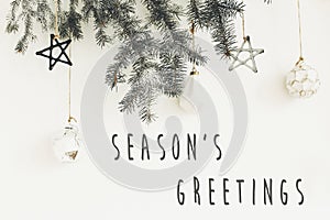 Season`s greetings text sign on stylish christmas branches with glass modern ornaments hanging on white wall. Creative christmas