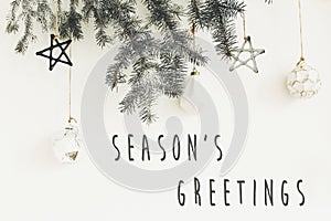 Season`s greetings text sign on stylish christmas branches with glass modern ornaments hanging on white wall. Creative christmas photo