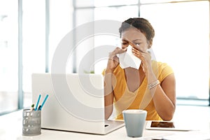 The season of the sniffles. a young businesswoman blowing her nose at her desk in a modern office.