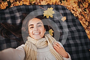 Season, technology and people concept - beautiful young woman lying on ground and autumn leaves and taking selfie with
