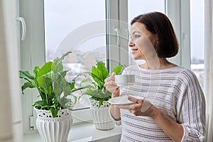 Season winter, snowy day, middle aged smiling woman with cup of coffee looking out the window.