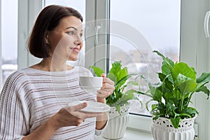 Season winter, snowy day, middle aged smiling woman with cup of coffee looking out the window.