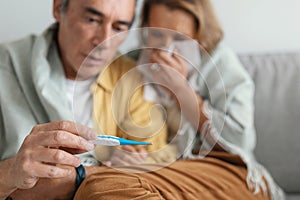 Seasonal flu. Worried senior man taking care of his ill wife at home, looking at thermometer with shock, selective focus