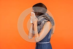 Seasonal influenza. Side view of flu-sick brunette woman wrapped in scarf, blowing her nose and sneezing in napkin