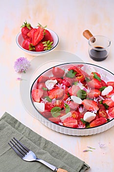 Seasonal salad with strawberries, tomatoes, soft cheese and chives flowers in a white plate on a light concrete background.