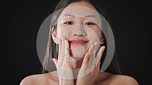 Seasonal skin moisturizing. Young beautiful asian woman applying nourishing cream on her cheeks, smiling to camera