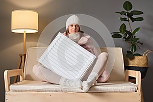 Seasonal temperature changes. Getting warm in winter. Calm woman wearing cap and coat sitting on couch and embracing heater