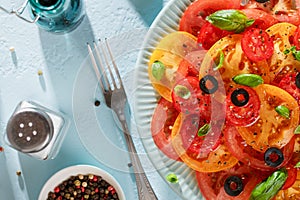 Seasonal tomato salad with basil, olives, spices and oil on blue table with fork and salt.