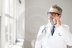 A seasoned doctor in a white coat is seen engaging in a phone conversation by a bright window