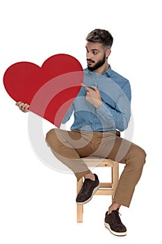 Seated curious man pointing his finger to a red heart