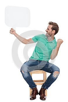 Seated sexy man celebrates victory while holding blank chat bubble