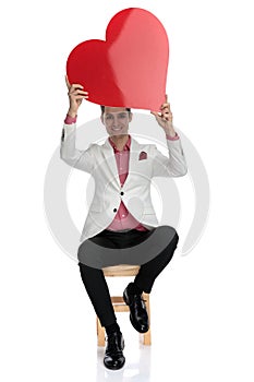 Seated smiling young businessman holds a red heart up