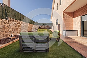 seating area of pallet sofas with green cushions next to a brick house with a garden and artificial grass floors