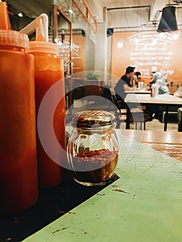 Sebuah botol kecil berisi cabe flakes di sebelah botol berisi saus sambal sebagai condiment di sebuah restoran. photo