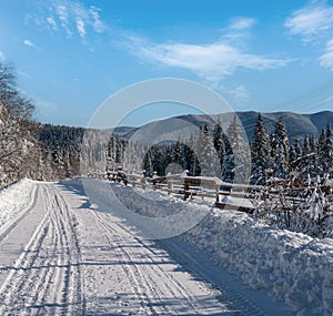 Secondary countryside alpine road to remote mountain hamlet through snowy fir forest, snow drifts and wood fence on wayside