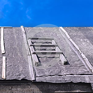 Section of a Wooden Ladder on a Barn Roof Covered with Waterproofing Tar Paper Against Summer Blue Sky. Roofing Repairs,
