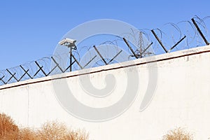 Security camera behind barbed wire fence stretched around prison walls. Security, surveillance camera watching area