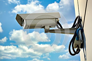 This is a security camera hanging on the wall of a building against a blue sky with clouds on a summer day.