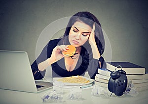 Sedentary lifestyle and junk food concept. Stressed woman sitting at desk eating hamburger