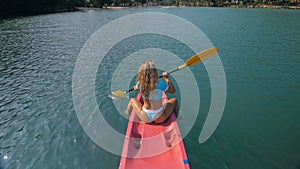 Seductive blonde woman in blue swimsuit sails on pink plastic kayak along azure sea at exotic resort backside upper