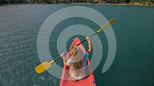 Seductive blonde woman in blue swimsuit sails on pink plastic kayak along azure sea at exotic resort backside upper