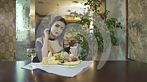 seductive brunet young woman in black dress with cleavage eating fruits grapes alone at table in fancy restaurant
