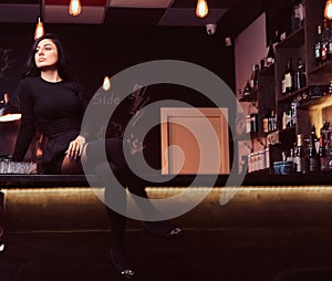 Seductive brunette girl in black clothes sitting with crossed legs on counter in a nightclub.