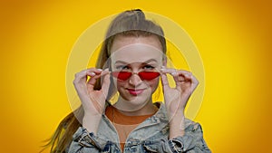 Seductive cheerful stylish girl in denim jacket wearing sunglasses, charming smile on yellow wall