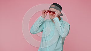 Seductive cheerful stylish man in blue shirt wearing sunglasses, charming smile on pink wall