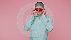 Seductive cheerful stylish man in blue shirt wearing sunglasses, charming smile on pink wall