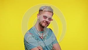 Seductive cheerful stylish man in blue t-shirt wearing sunglasses, charming smile on yellow wall
