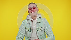 Seductive cheerful stylish man in denim jacket wearing sunglasses, charming smile on yellow wall