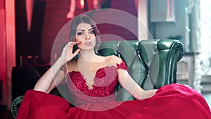 Seductive Hispanic young girl in glamour red evening dress sitting on vintage armchair medium shot