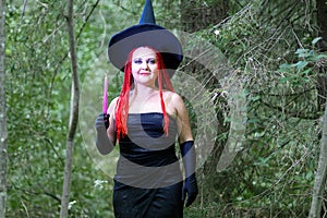 Seductive sorceress with red hair in a pointed hat in the forest is engaged in magic with a candle.