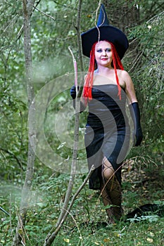 Seductive sorceress with red hair in a pointed hat in the forest is engaged in magic with a candle.