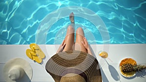 Seductive woman in hat sitting at poolside, enjoying summer time, vacation