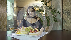 seductive young brunet woman in black dress with cleavage eating fruits grapes alone at table in fancy restaurant