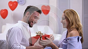 Seductive young female giving red gift box to surprised boyfriend, anniversary